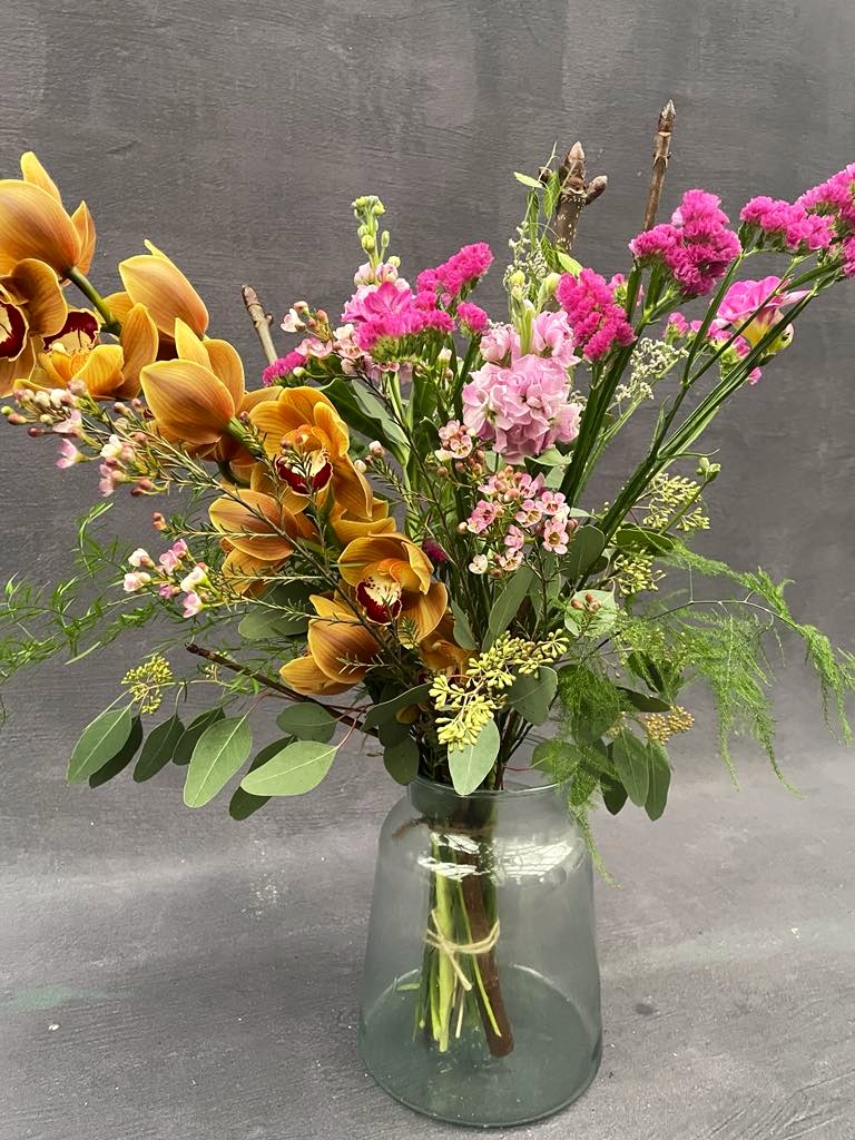 Høj og luftig buket med blomster i orange og pink farver, står i en glasvase. 