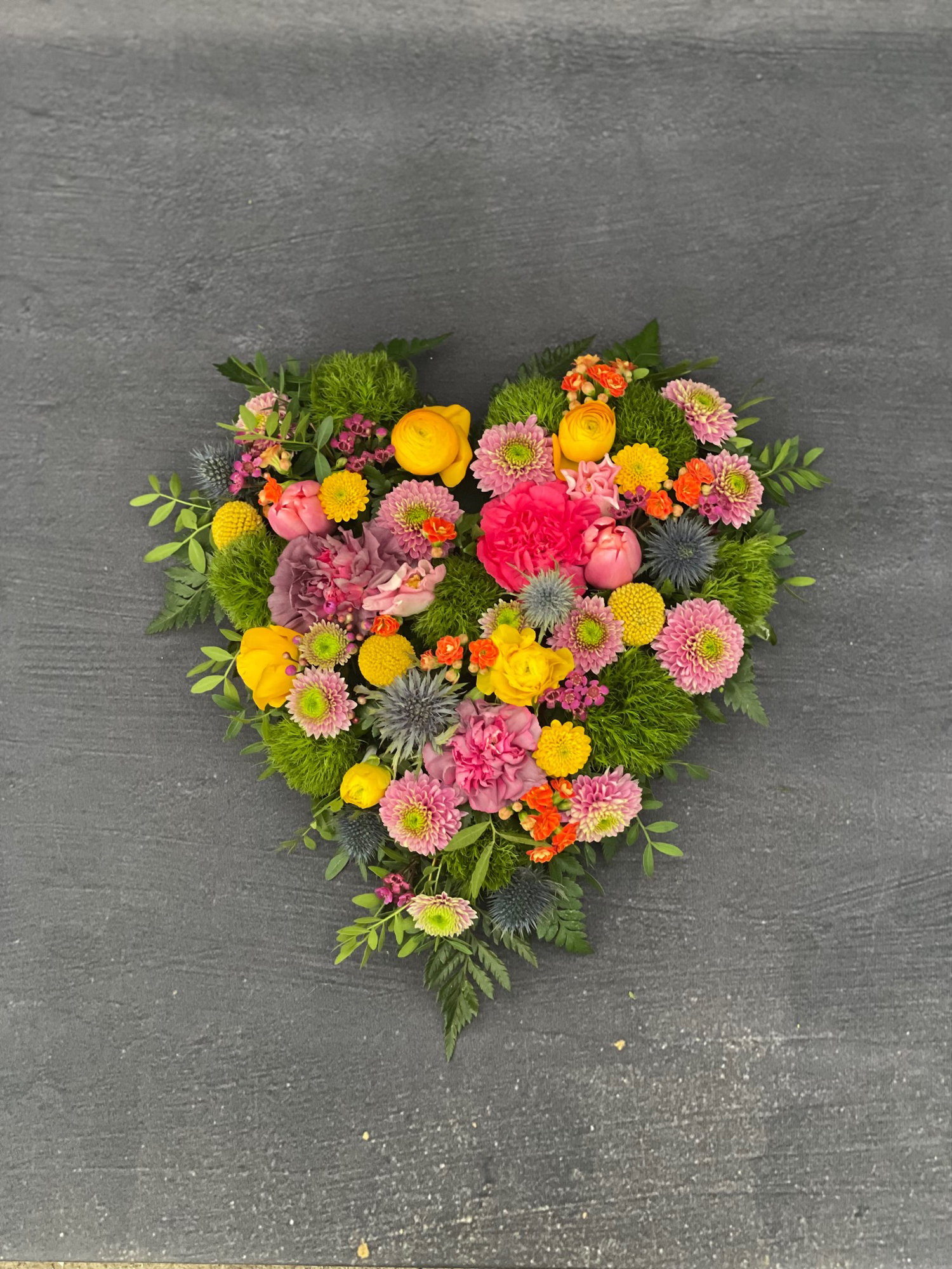 Oasis hjerte til begravelse. Hjertet er bundet med blomster i pink, gule og orange nuancer. 