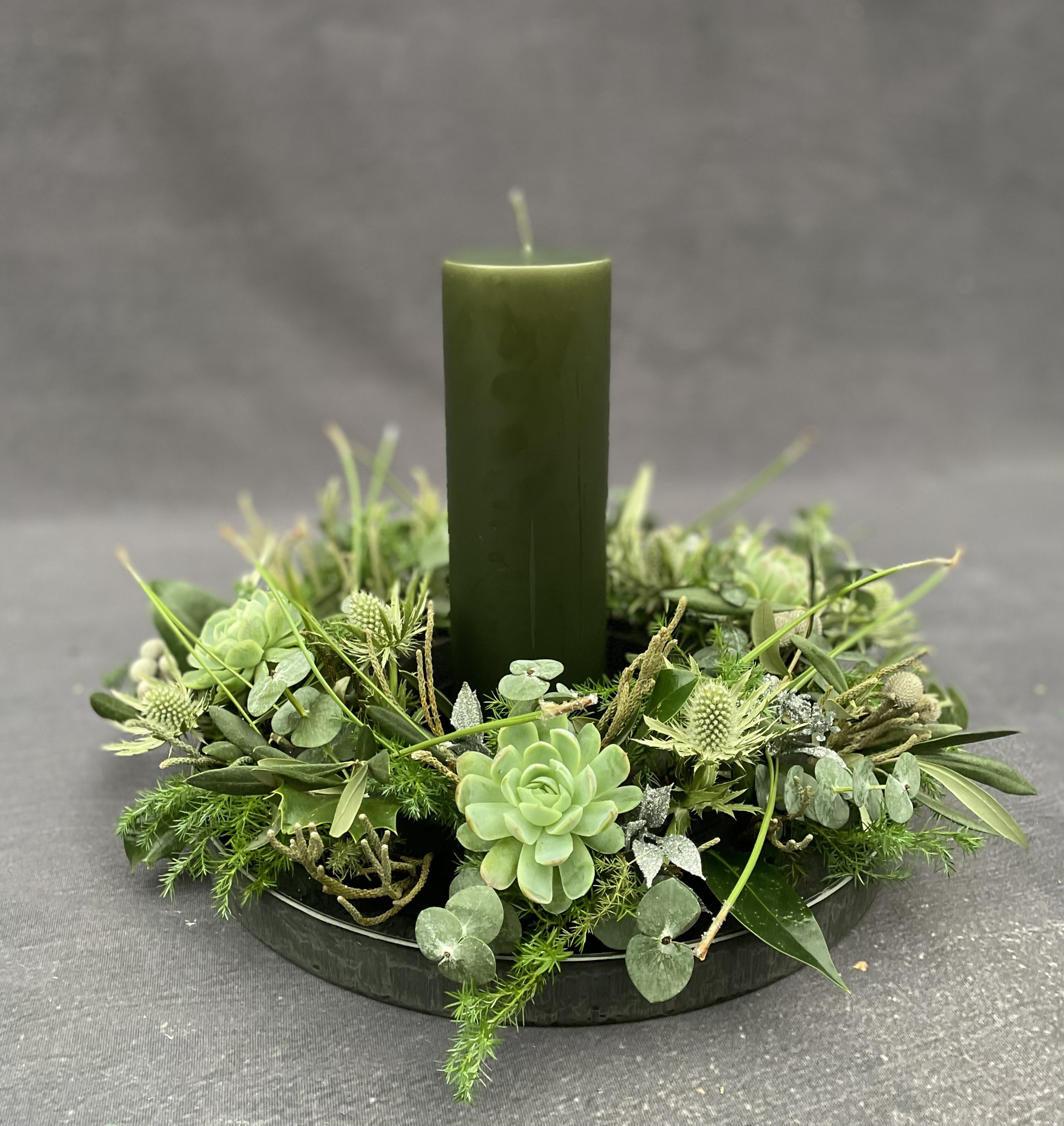 Grøn krans bundet med husløg, mandstro, eukalyptus og grønne græsser. I midten står et grønt bloklys. 