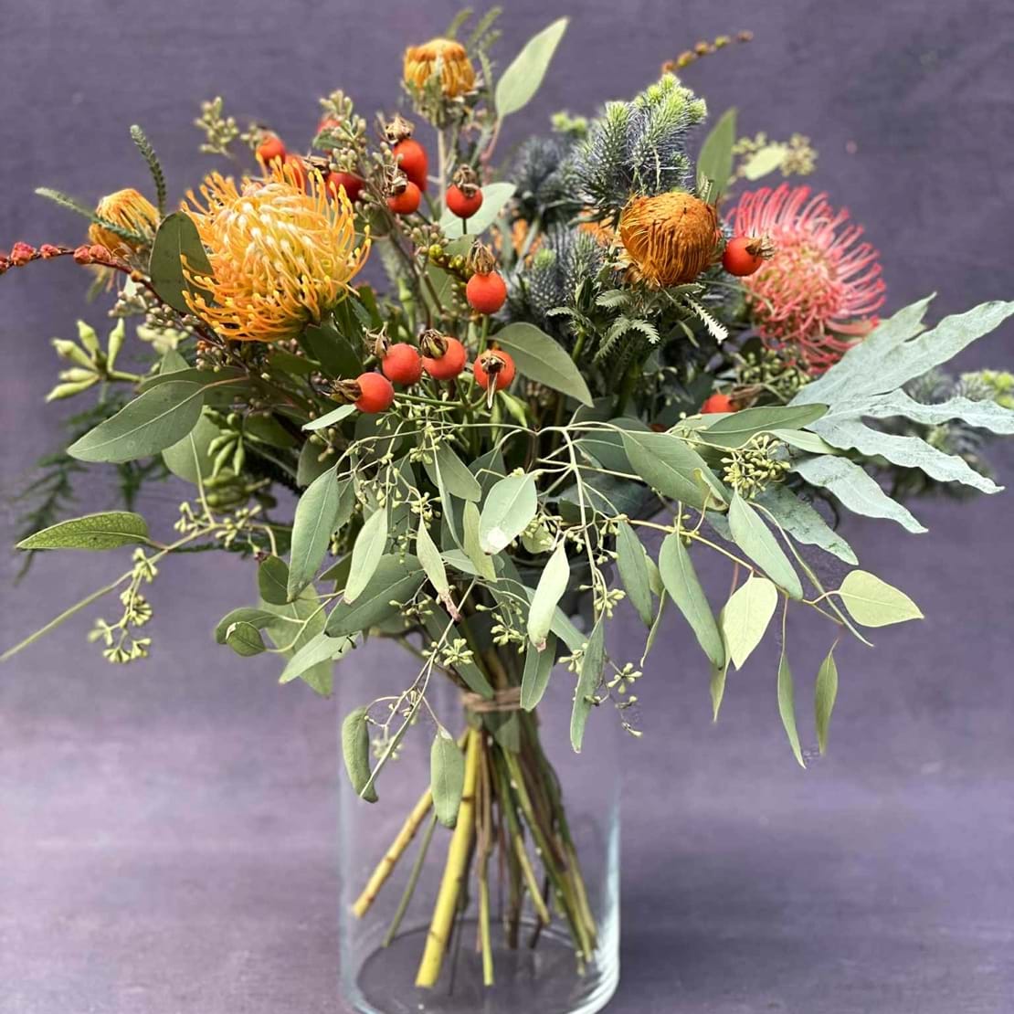 Efterårsbuket i klarglasvase. Buketten er bundet i et vildt udtryk, med blomster i varme orange og røde nuancer, samt grønt.