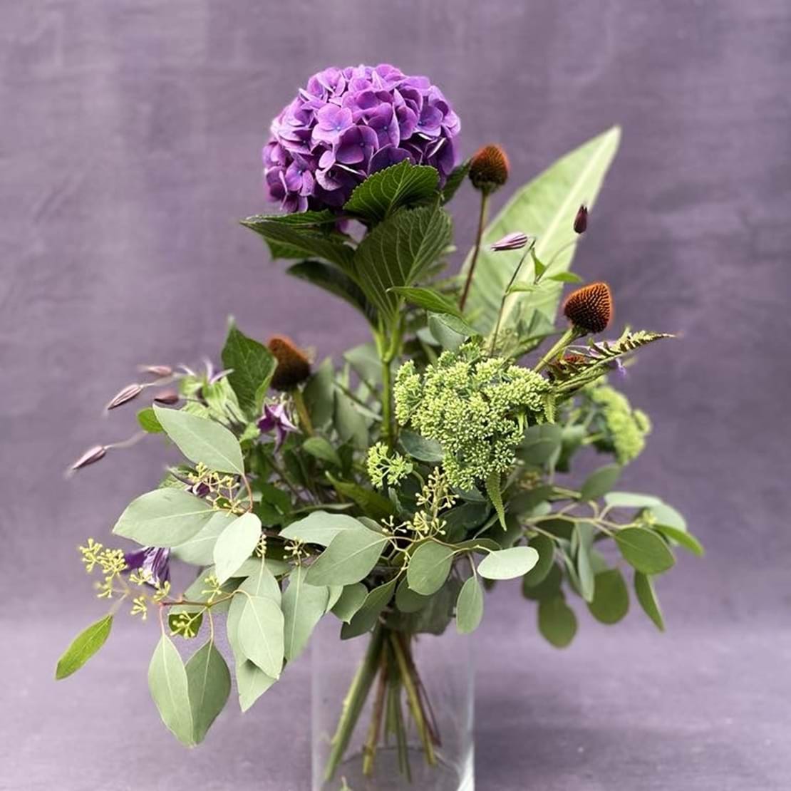 Høj og luftig buket med et livligt udtryk af en stor lilla hortensia og lysegrønne sommerblomster.