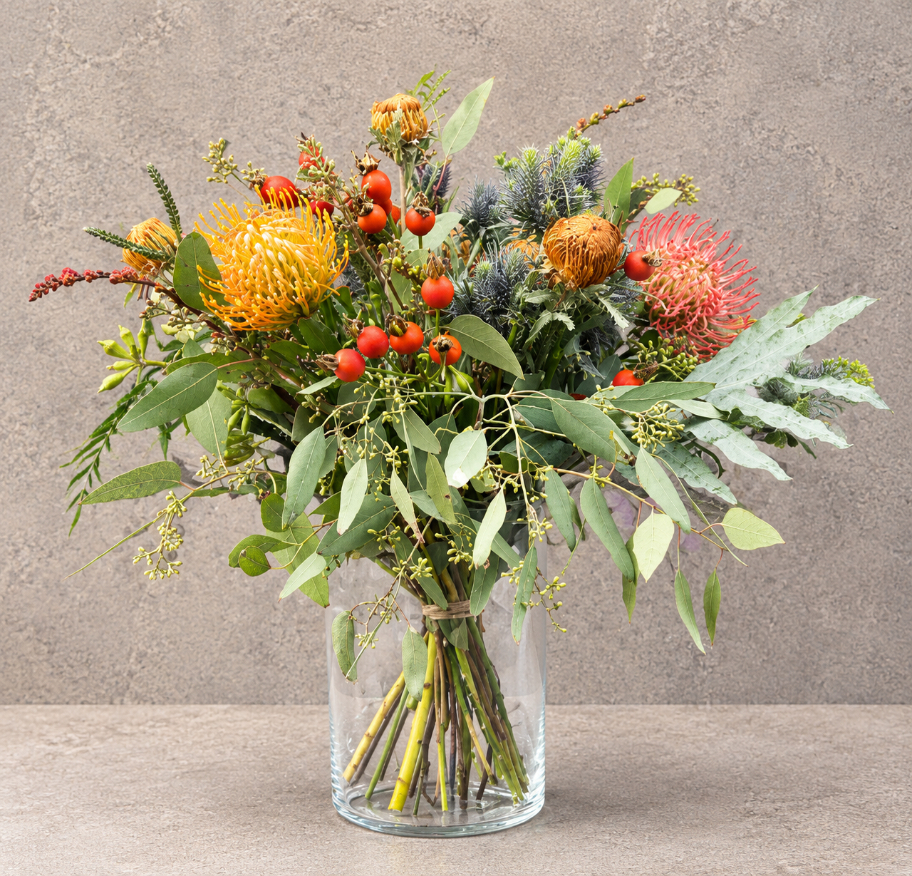 Efterårsbuket i klarglasvase. Buketten er bundet i et vildt udtryk, med blomster i varme orange og røde nuancer, samt grønt.