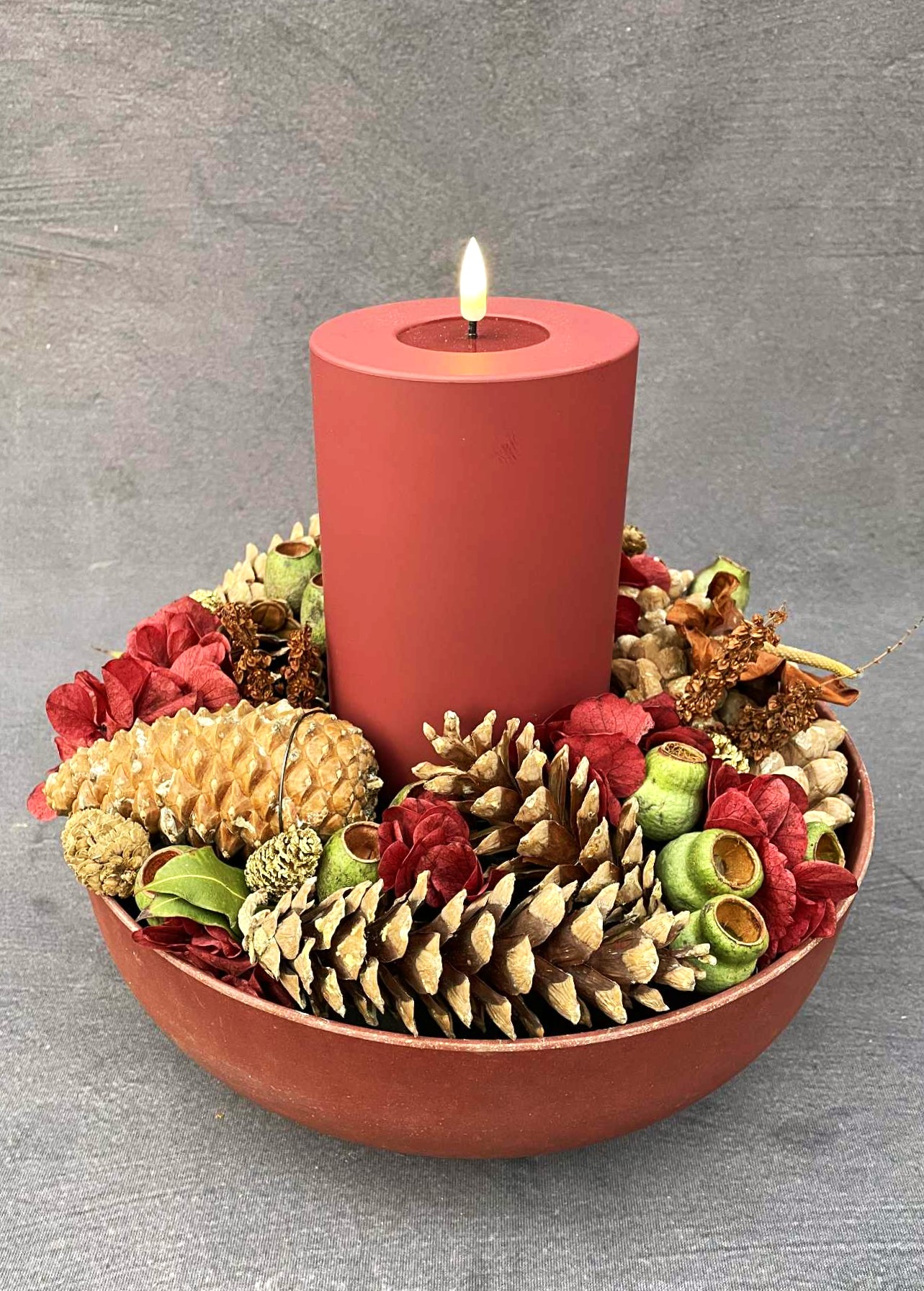 Juledekoration med rundt fad fyldt af kogler, agern og tørrede blomster. På toppen lyser et rødt LED bloklys.